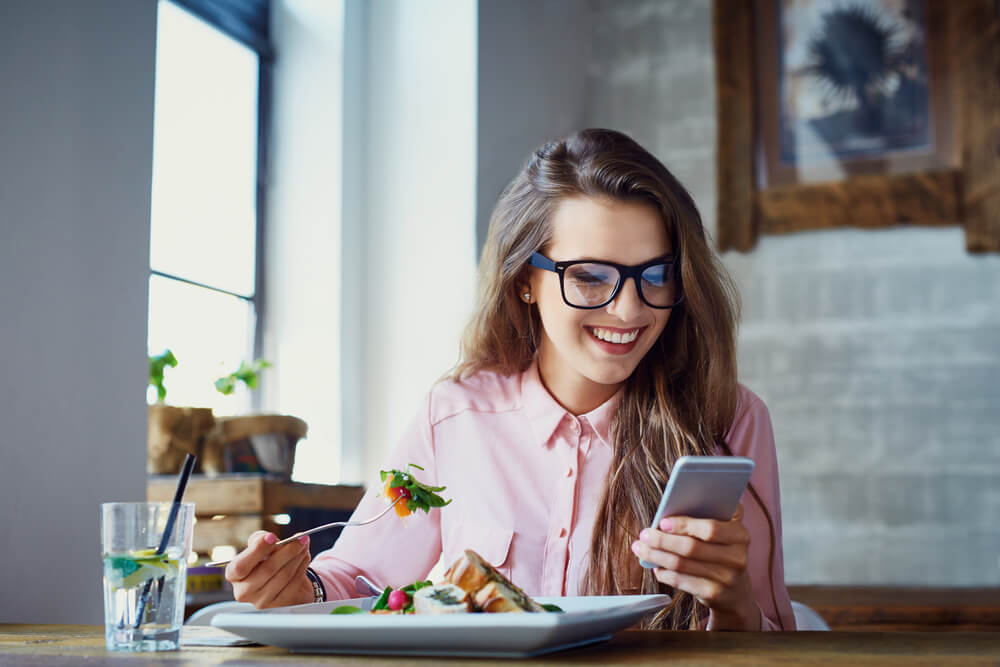 Smiling young woman eating healthy salad while texting on the phone
