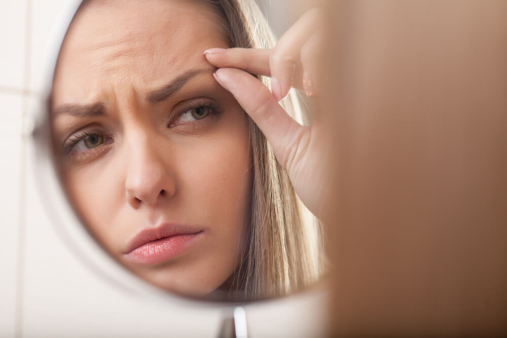 Woman frowning while examining forehead wrinkles in a dressing mirror